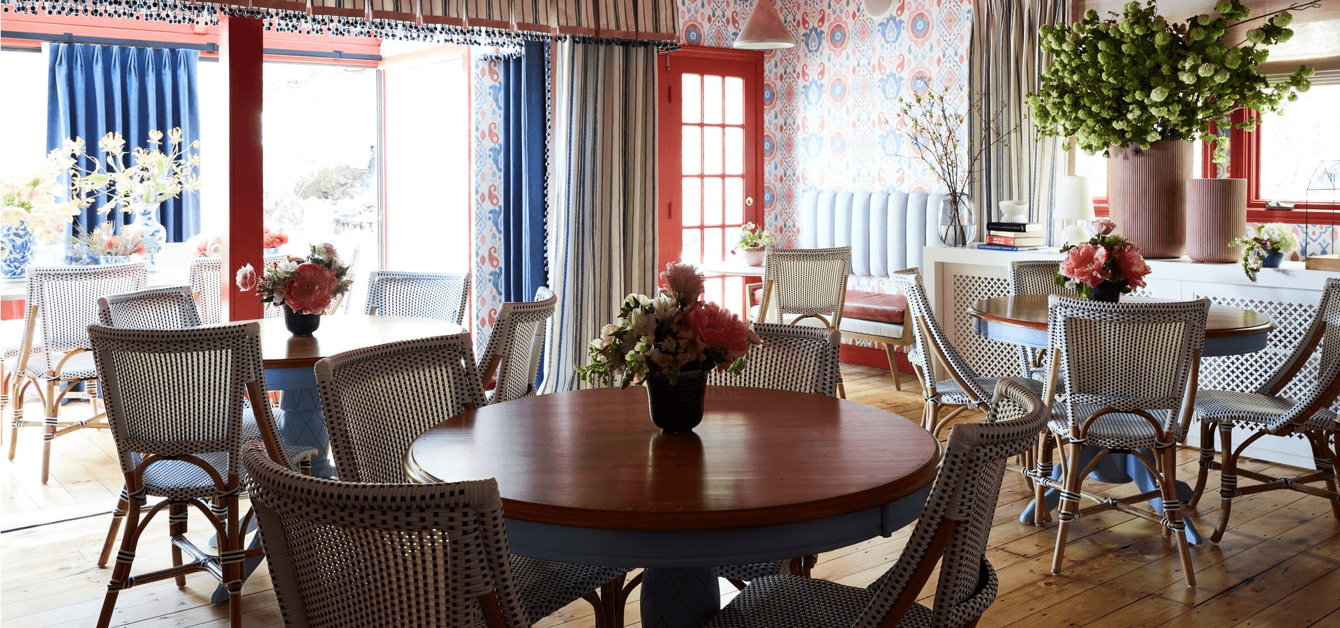 Brightly decorated dining area featuring wooden tables, rattan chairs, and floral arrangements against a colorful backdrop.