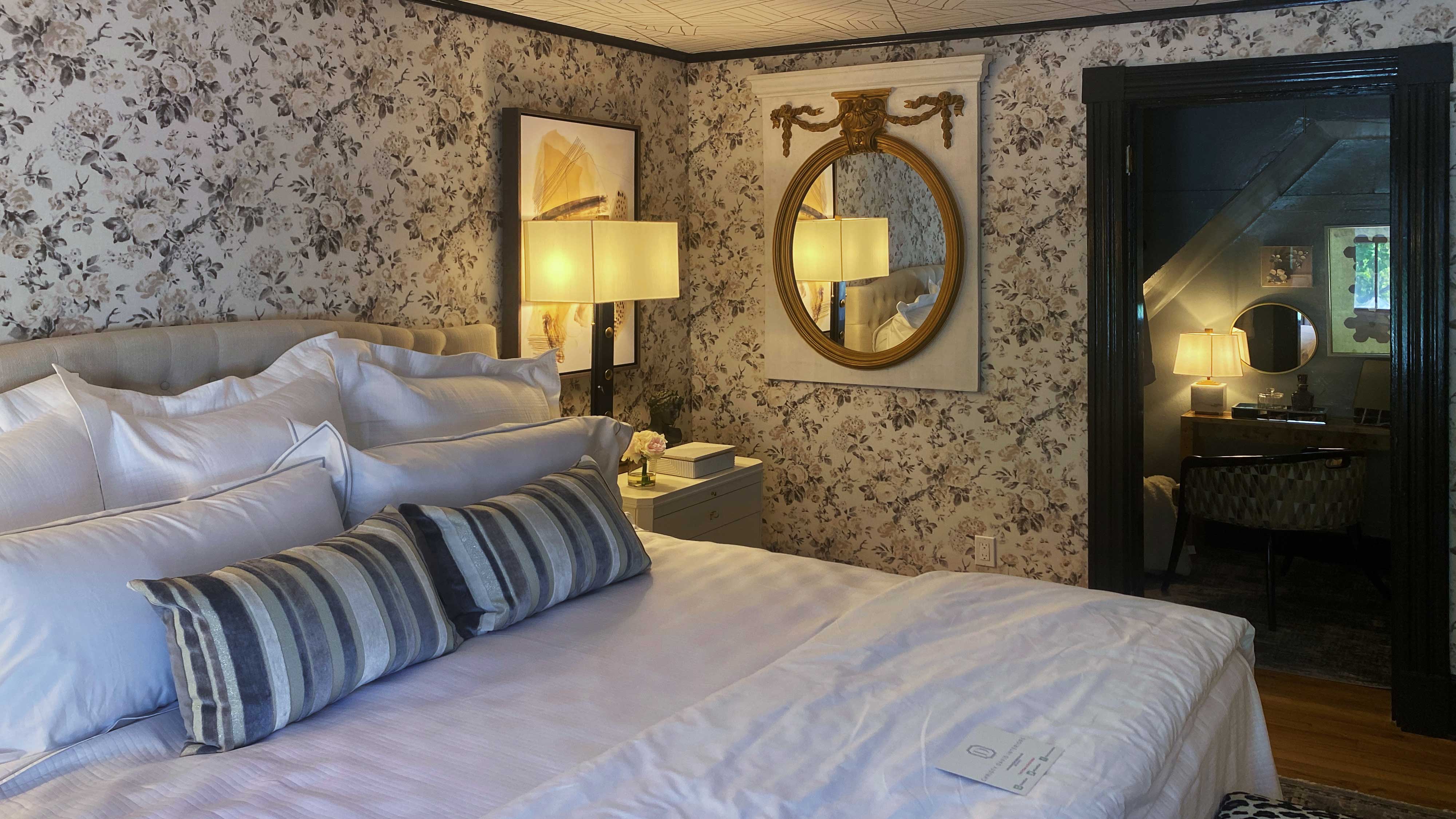 A bedroom with floral wallpaper, a made bed with pillows, a framed mirror, and a doorway leading to another room.