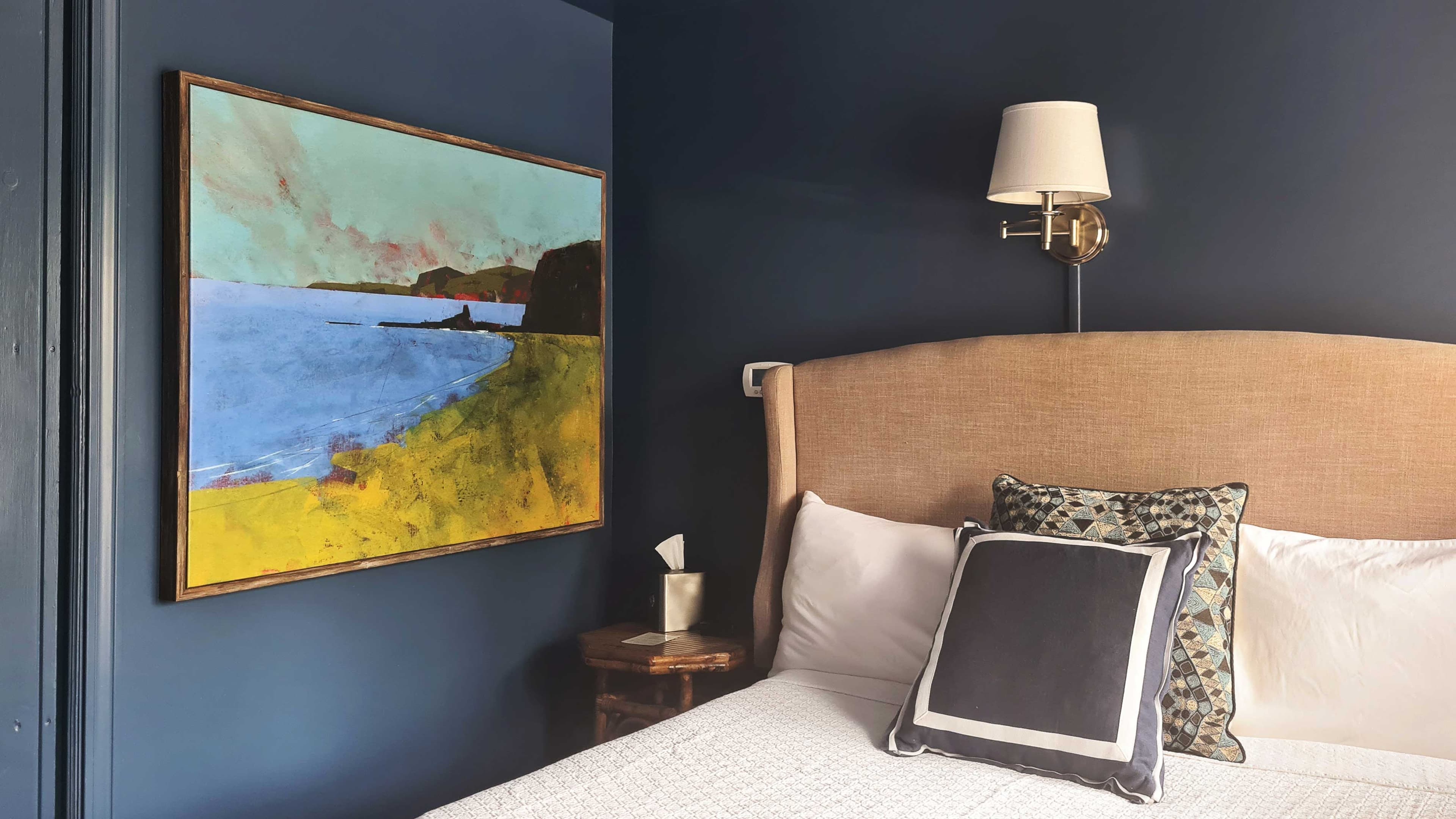 A bedroom with blue walls, a painting, and a bed with pillows.
