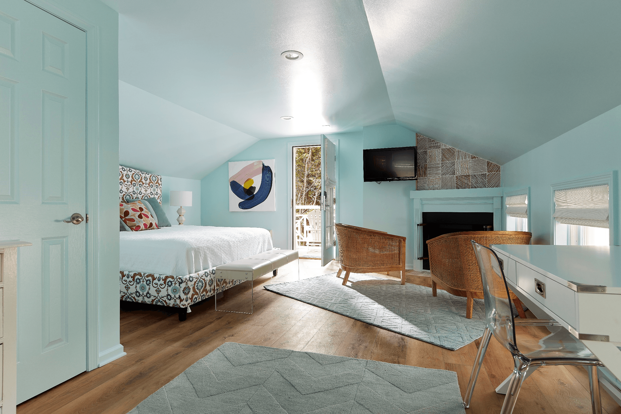 A light blue bedroom with a bed, two wicker chairs, a desk, and a door leading outside.