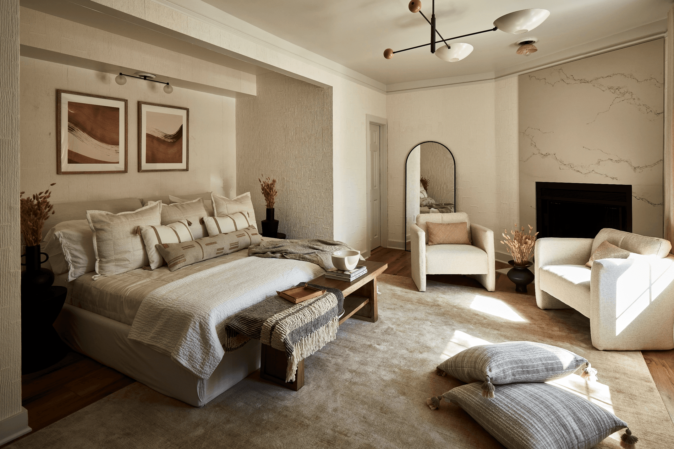 A neutral-toned bedroom with a made bed, two armchairs, a fireplace, and artwork.