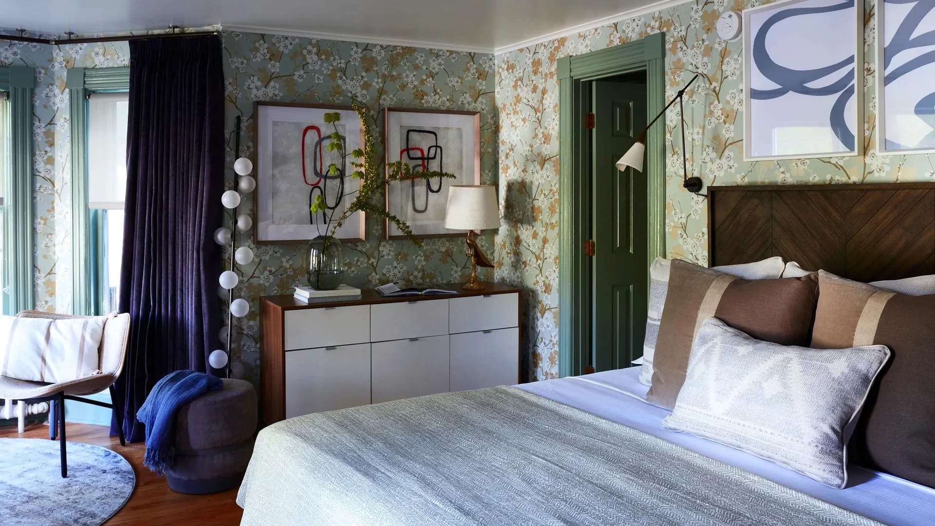 vibrant room with green print wallpaper, wooden modern dresser with artwork hung above and bed with throw pillows