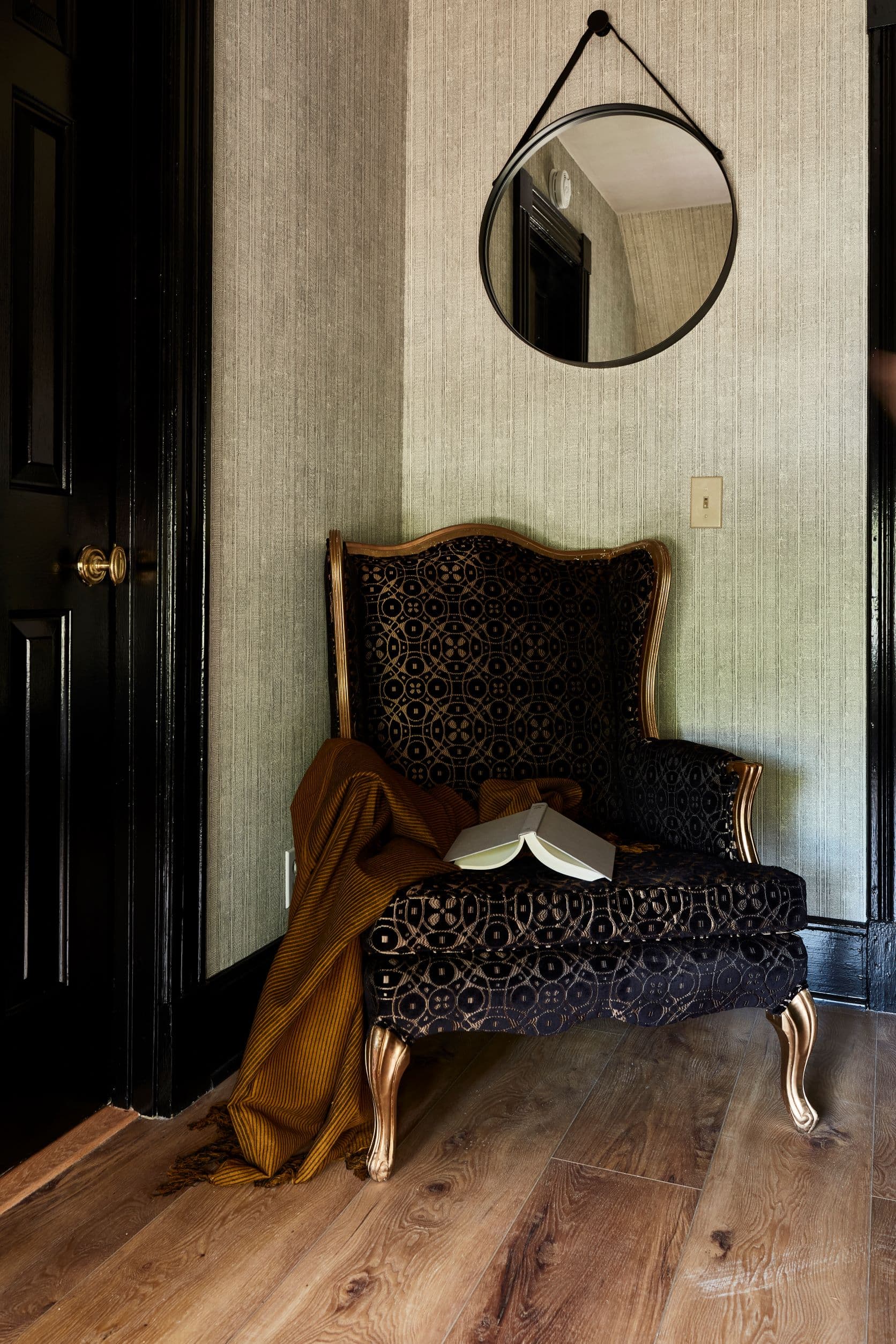 A patterned armchair with a book and throw blanket, a round mirror, and a dark doorway.