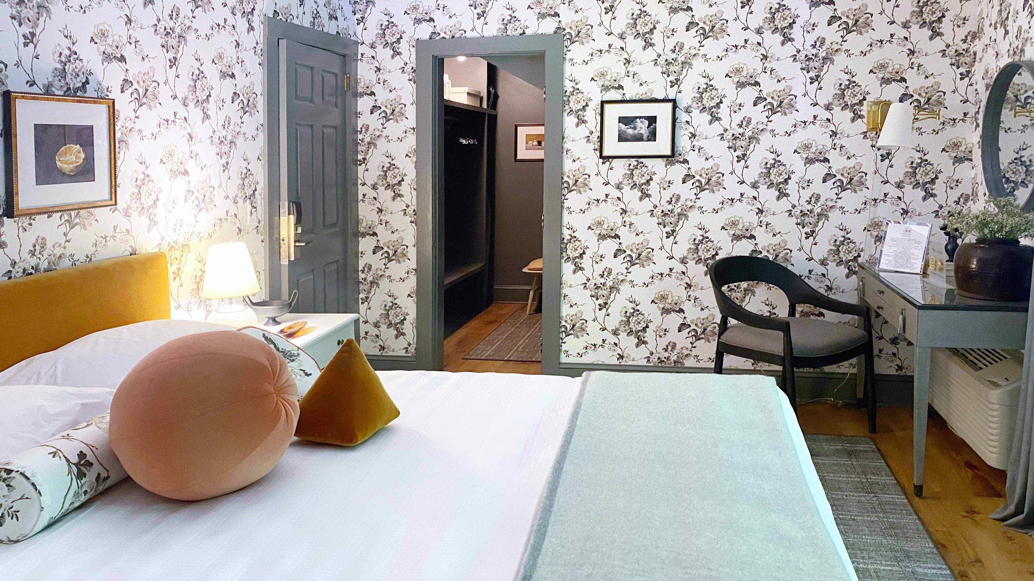 A bedroom with floral wallpaper, a bed, a door leading to another room, a chair, and a desk.