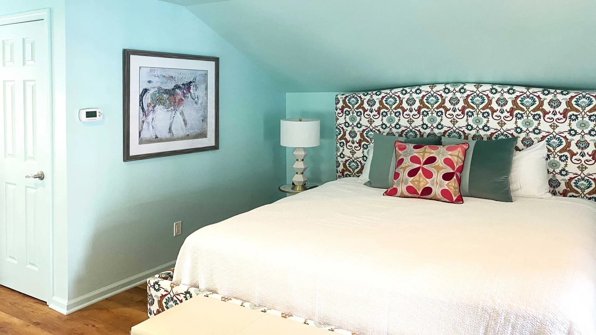 Blue bedroom with patterned headboard, framed art, and a small lamp.