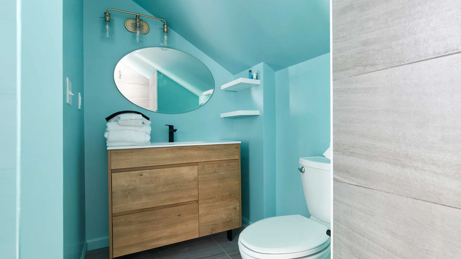 Small, modern bathroom with blue walls, round mirror, and wood vanity.