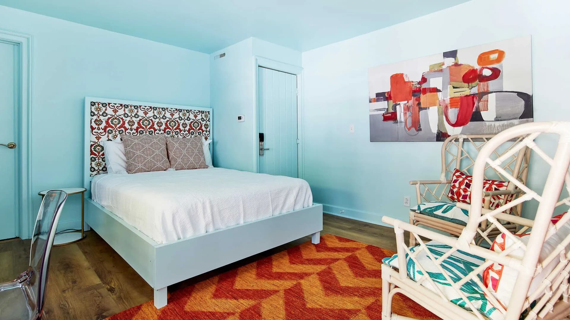 Blue bedroom with patterned headboard, orange rug, and two wicker chairs.