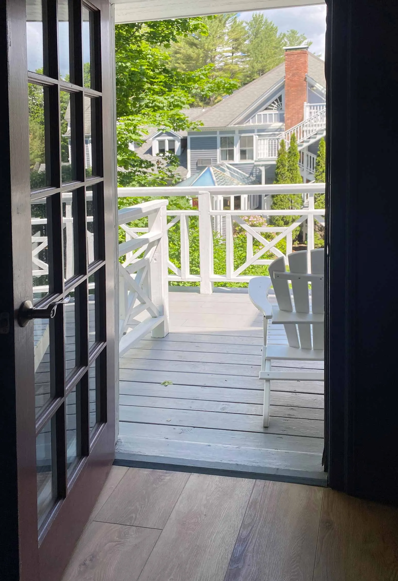 View from inside a room through open French doors to a balcony with white railings and a white Adirondack chair.
