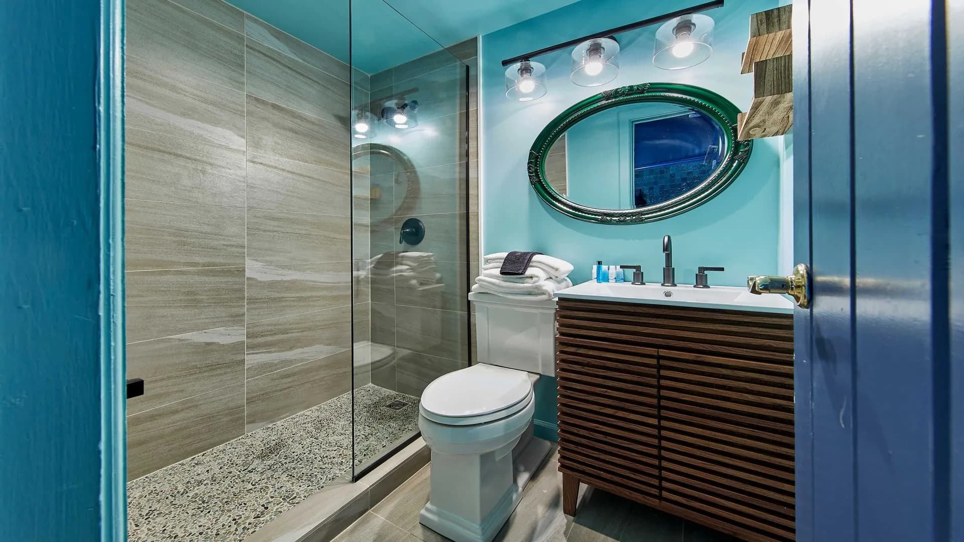 Modern blue bathroom with glass shower, unique vanity, and round mirror.