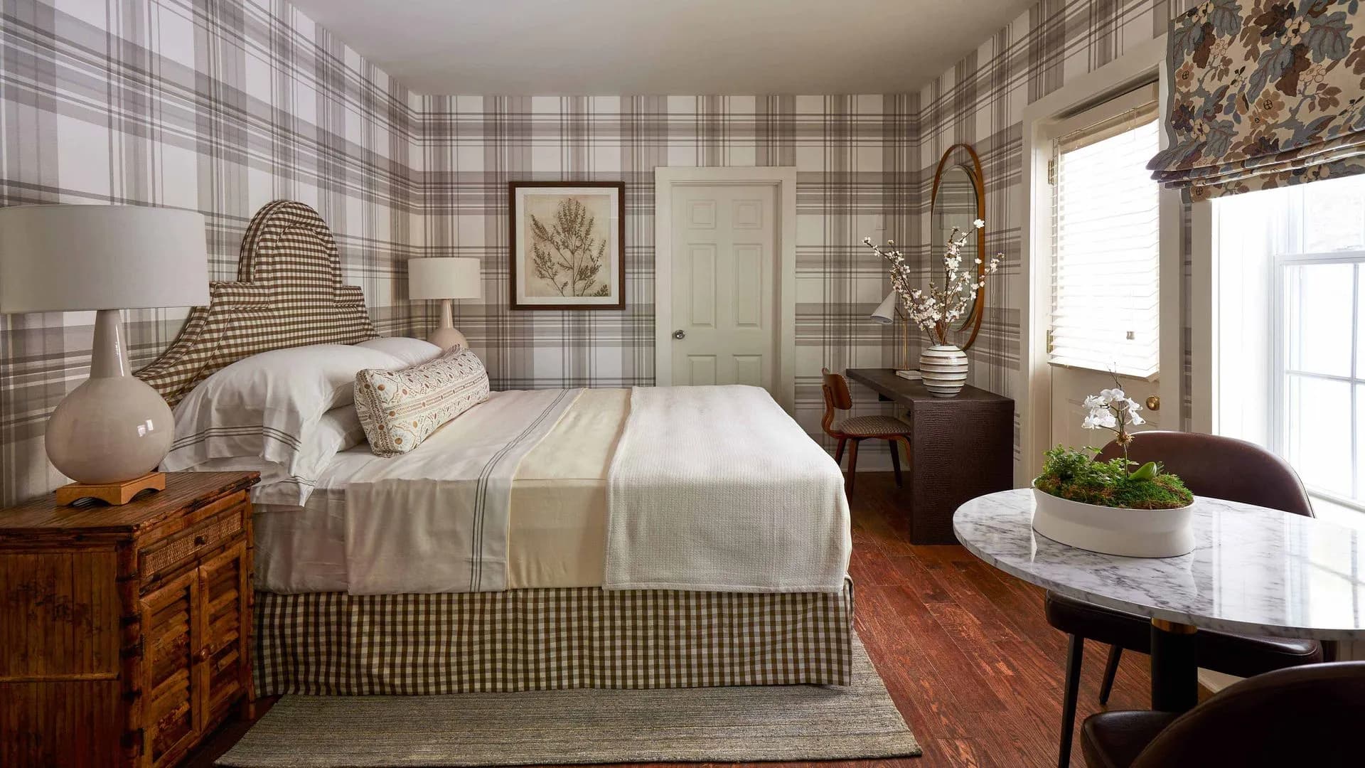 Light brown and white checkered wallpaper with tan bedframe and white sheets with a small table and chair