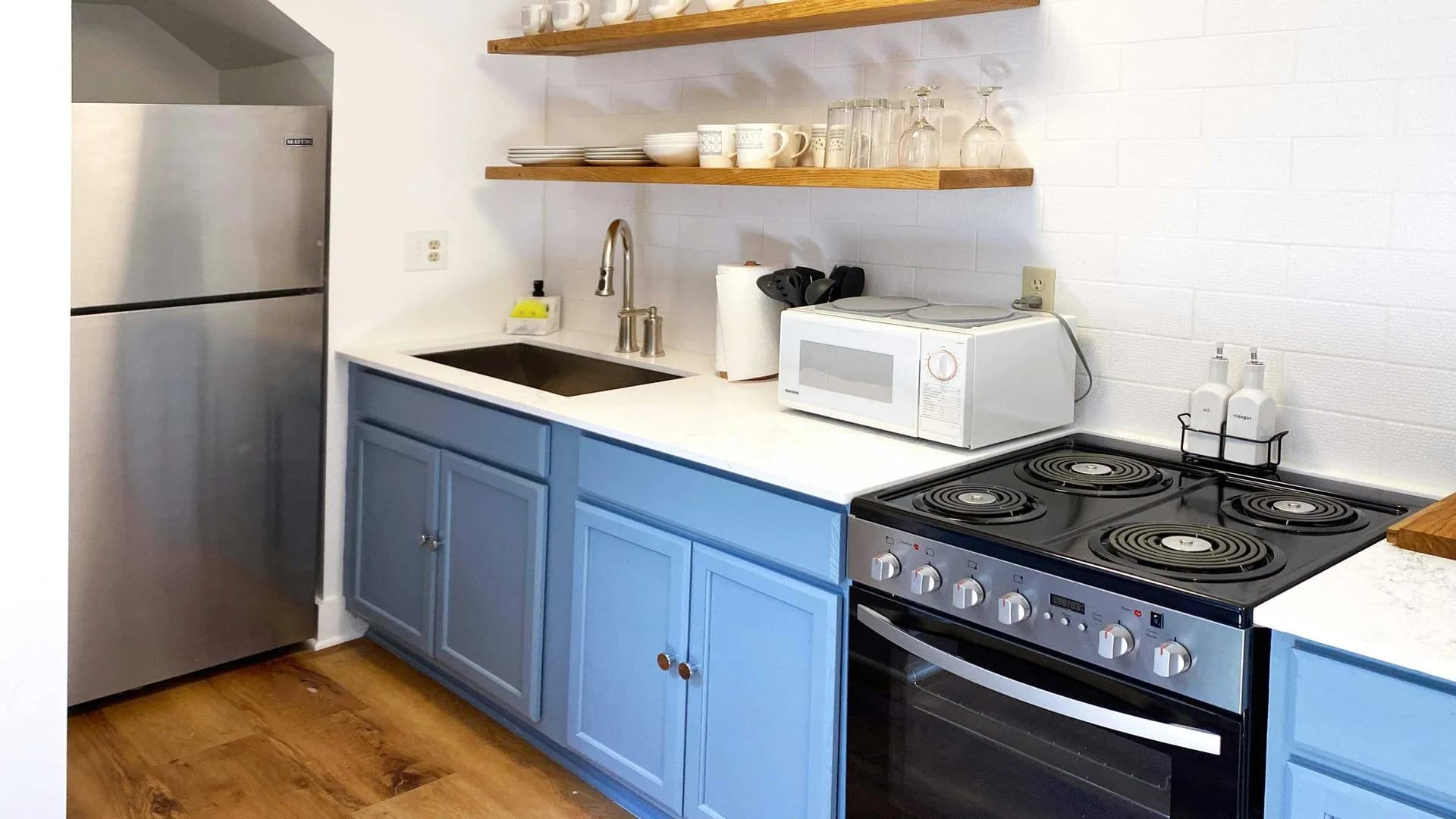 Small modern kitchen with blue cabinets, open shelving, and white tile backsplash.