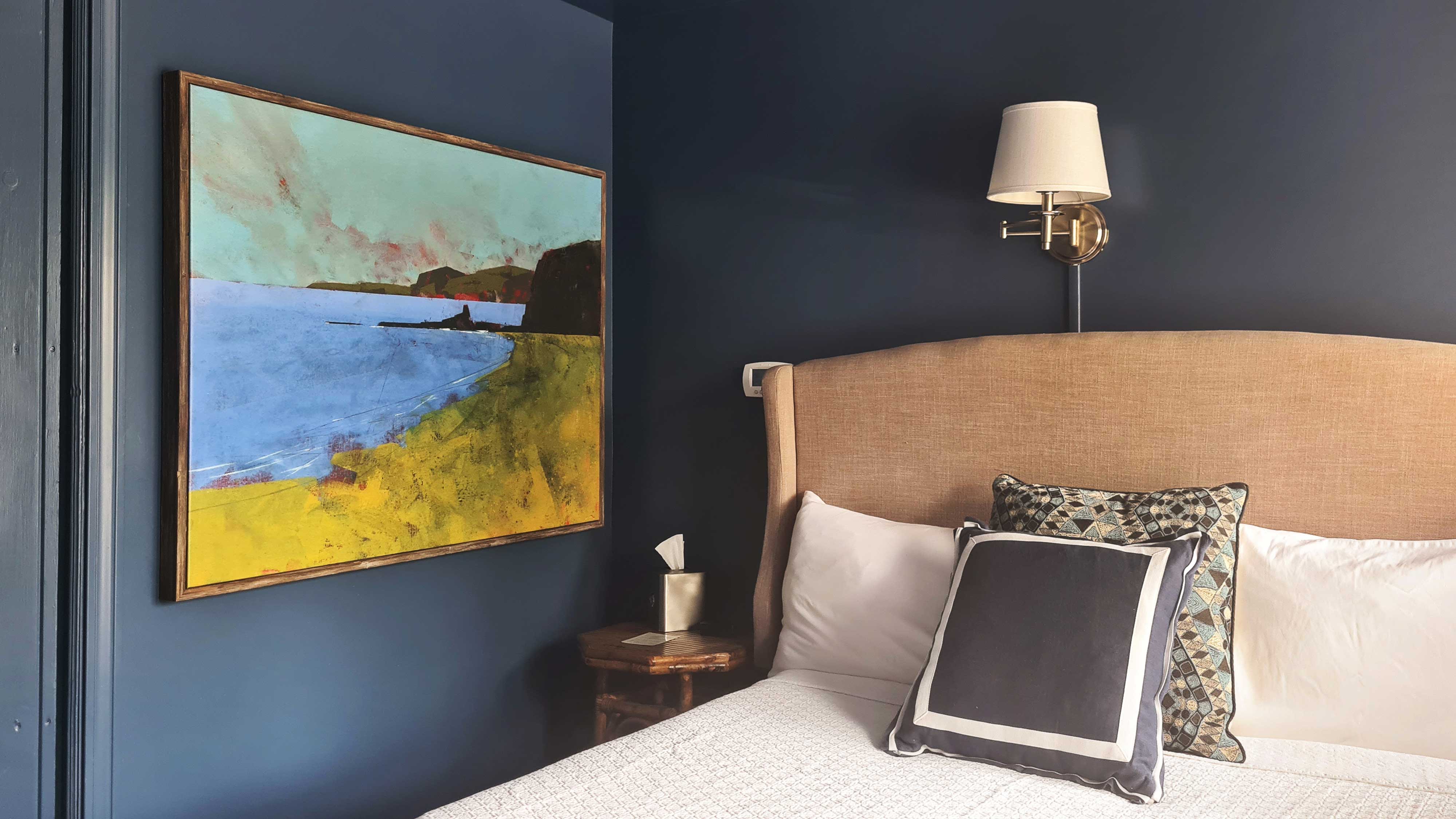 A bedroom with blue walls, a painting, and a bed with pillows.