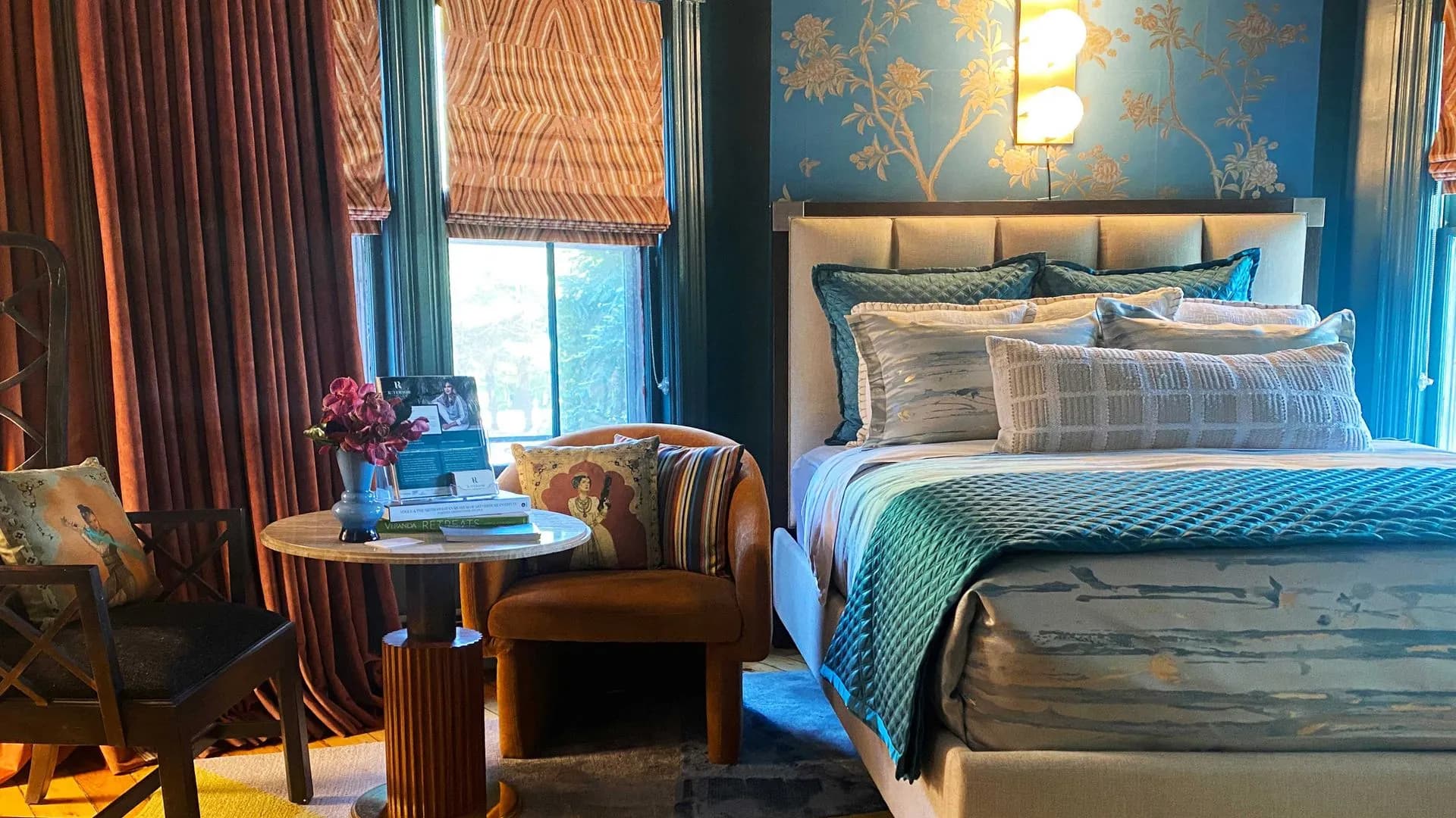 Elegant bedroom with a plush bed, decorative pillows, rich blue and gold wallpaper, and cozy seating area.