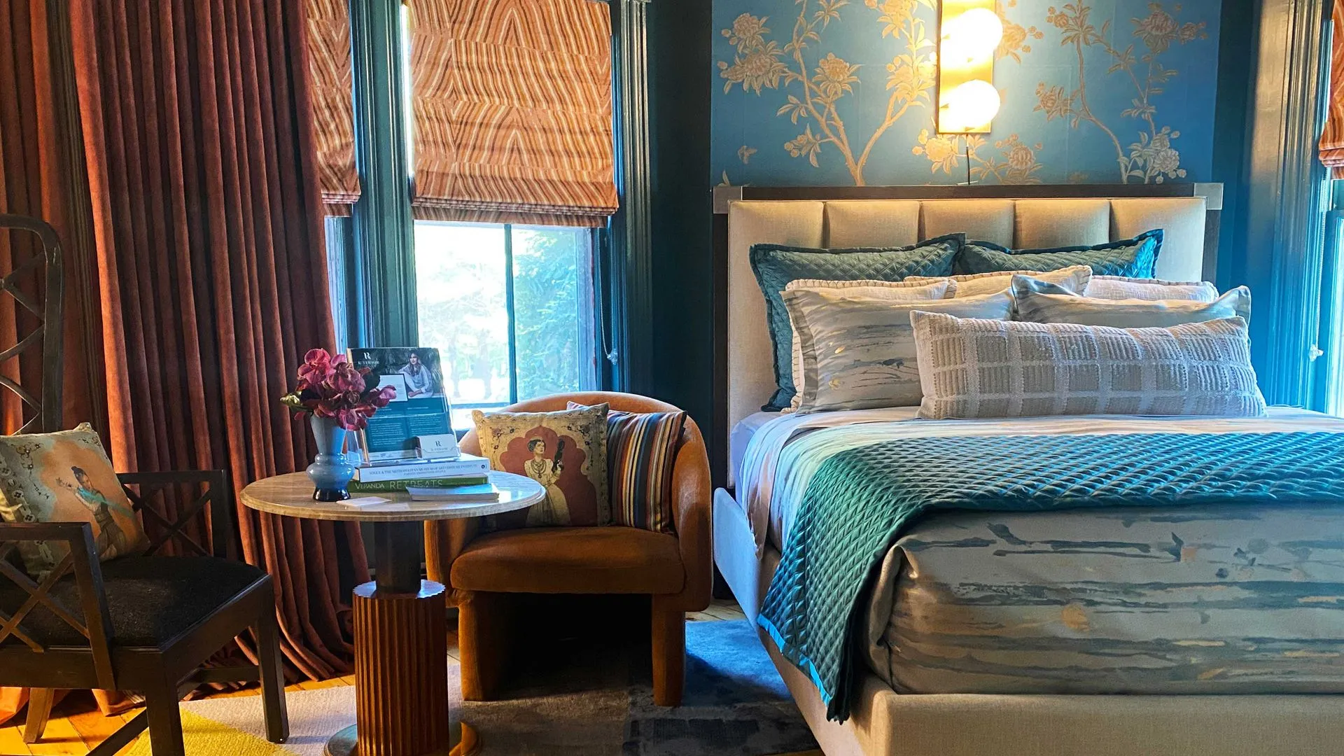 Elegant bedroom with a plush bed, decorative pillows, rich blue and gold wallpaper, and cozy seating area.